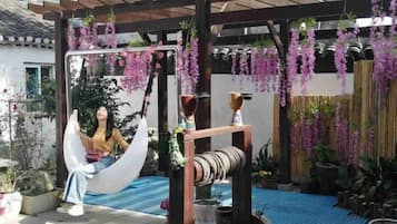 A hammock under a pergola with hanging purple flowers, surrounded by potted plants and a blue mat.