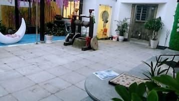 Ein Innenhof mit einem mondsichelförmigen Pflanzgefäß, Topfpflanzen, einem Tisch mit Unterlage und einer traditionellen Wasserpumpe.