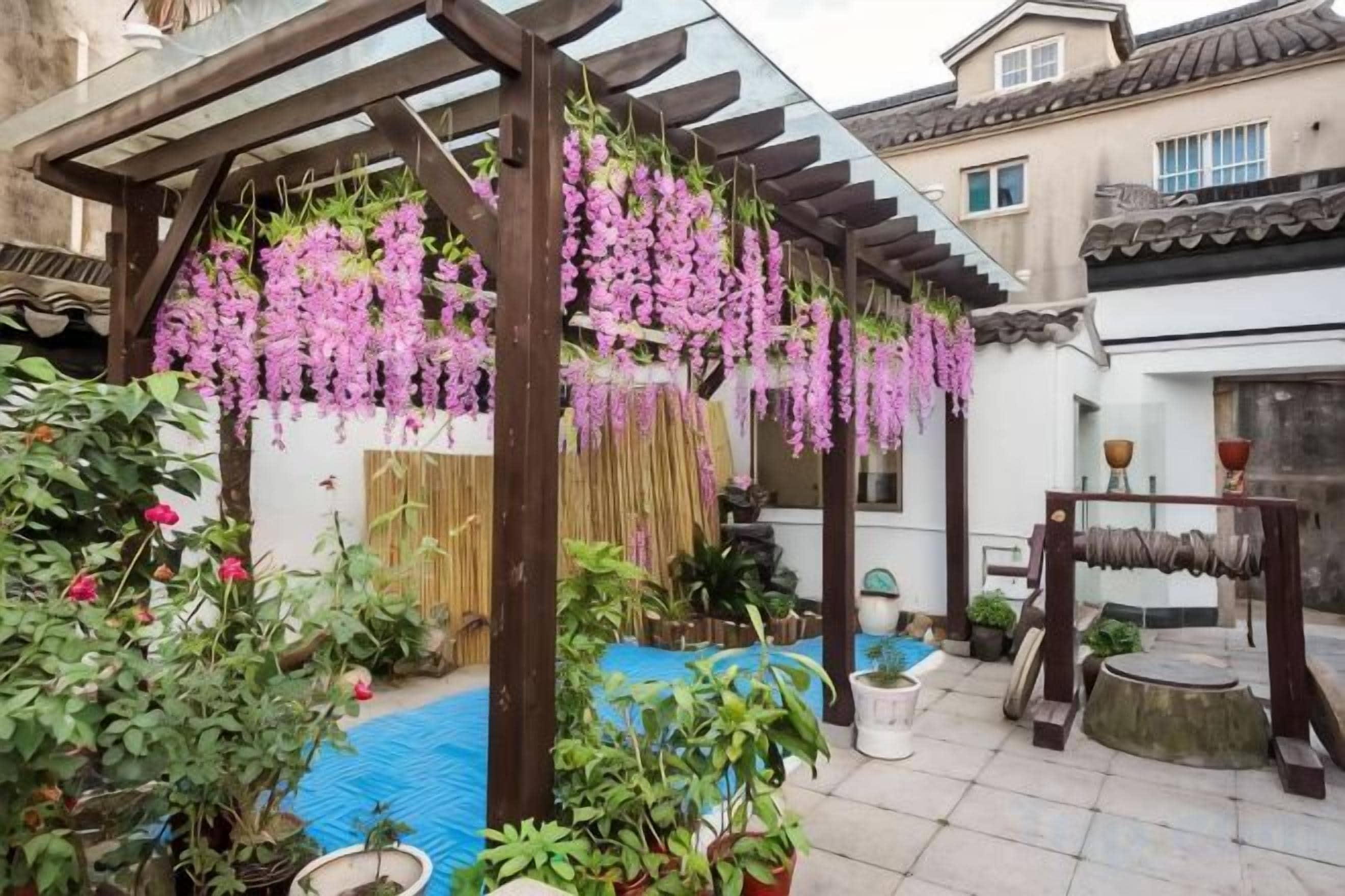 A covered outdoor area with hanging purple flowers, potted plants, and a wooden structure.