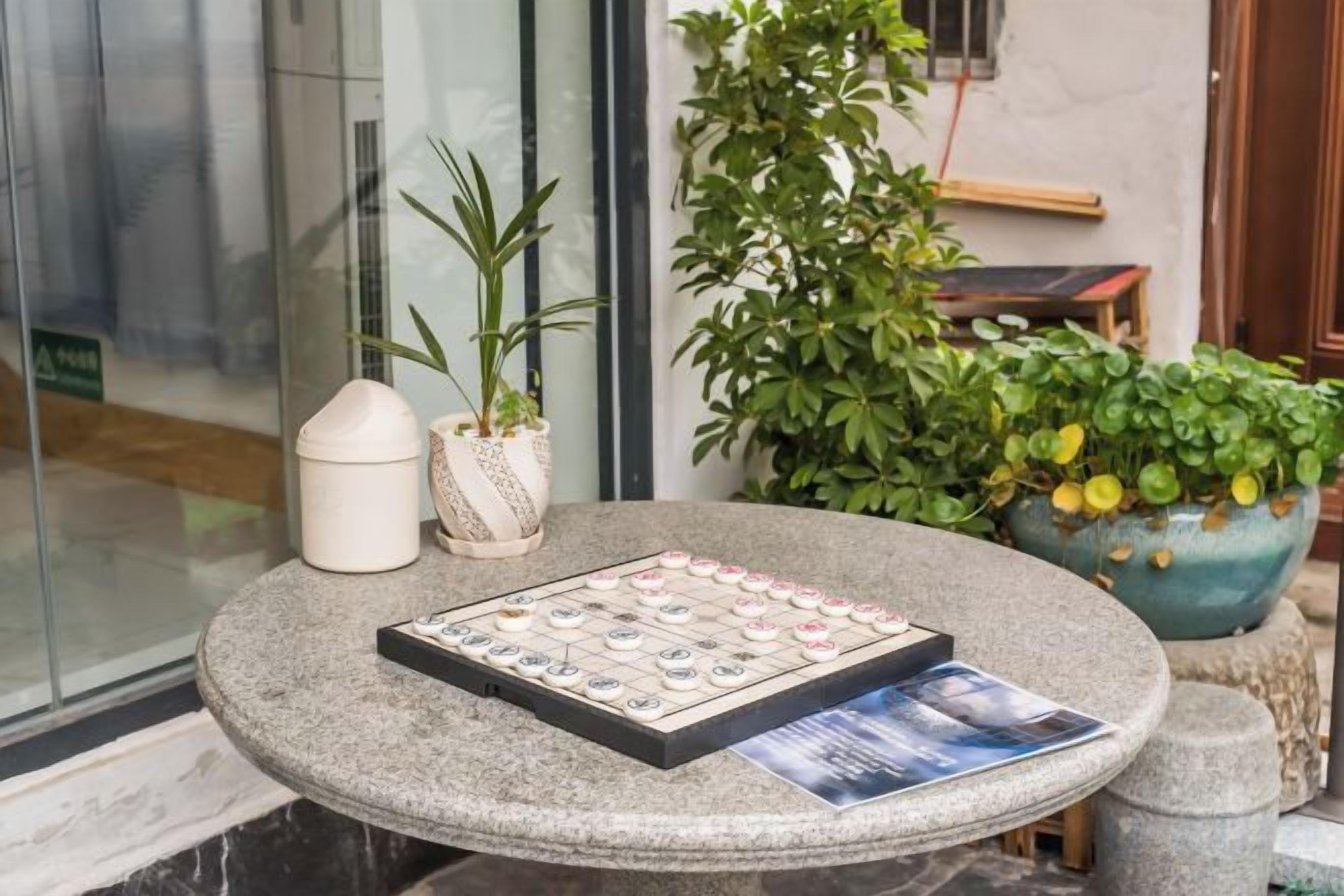 A round stone table with a chessboard and a magazine, surrounded by potted plants.