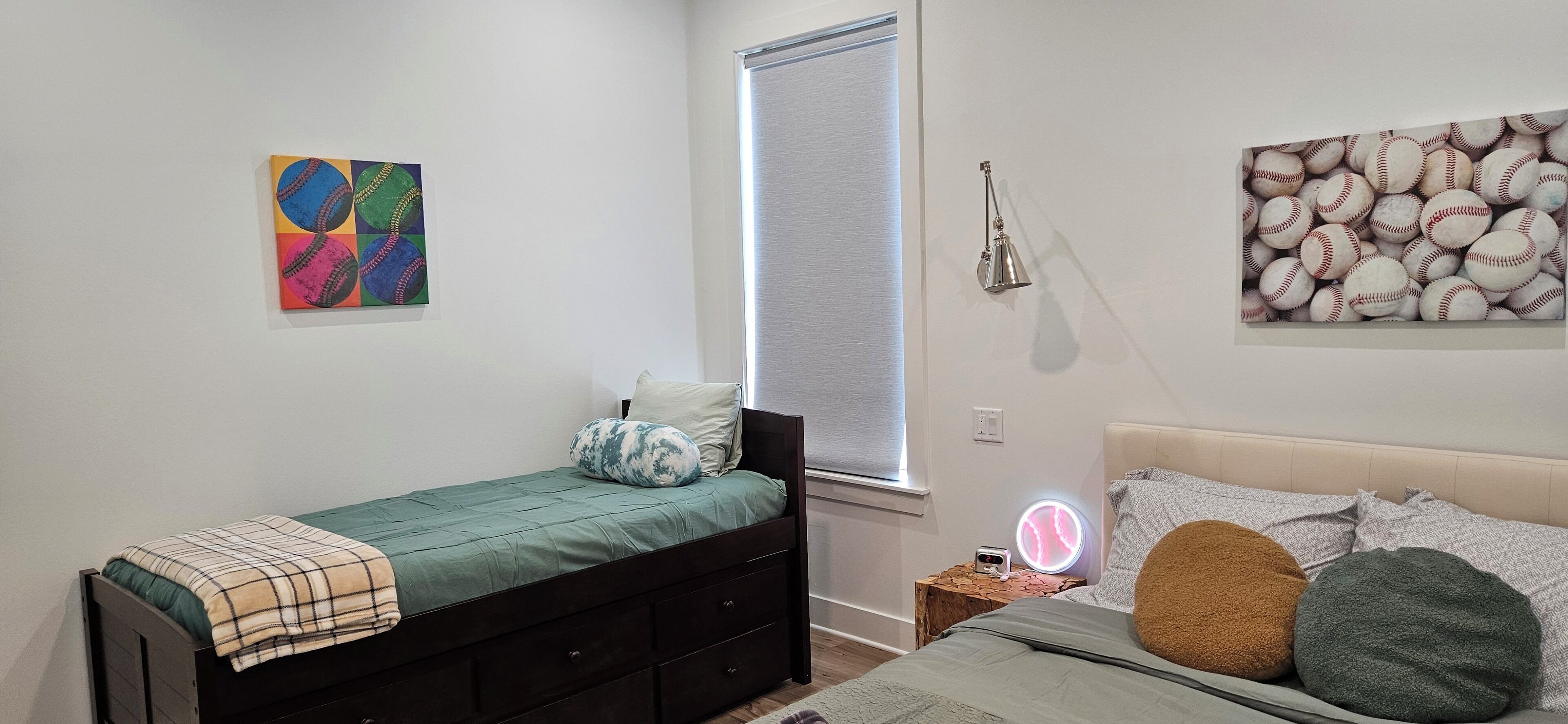 A bedroom with a single bed, a bedside table with a lamp, a wall art of baseballs, and a window with blinds.