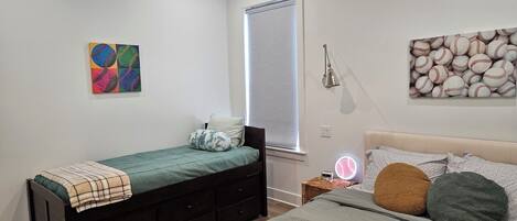 A bedroom with a single bed, a bedside table with a lamp, a wall art of baseballs, and a window with blinds.