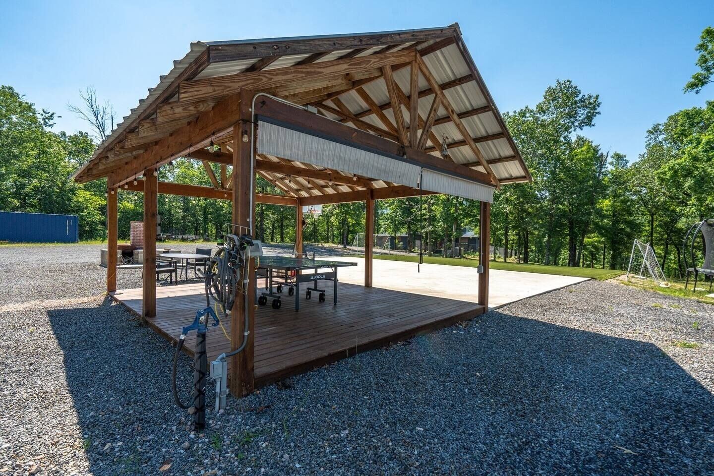 A covered wooden pavilion with a table and chairs, a ping pong table, and a metal swing set.