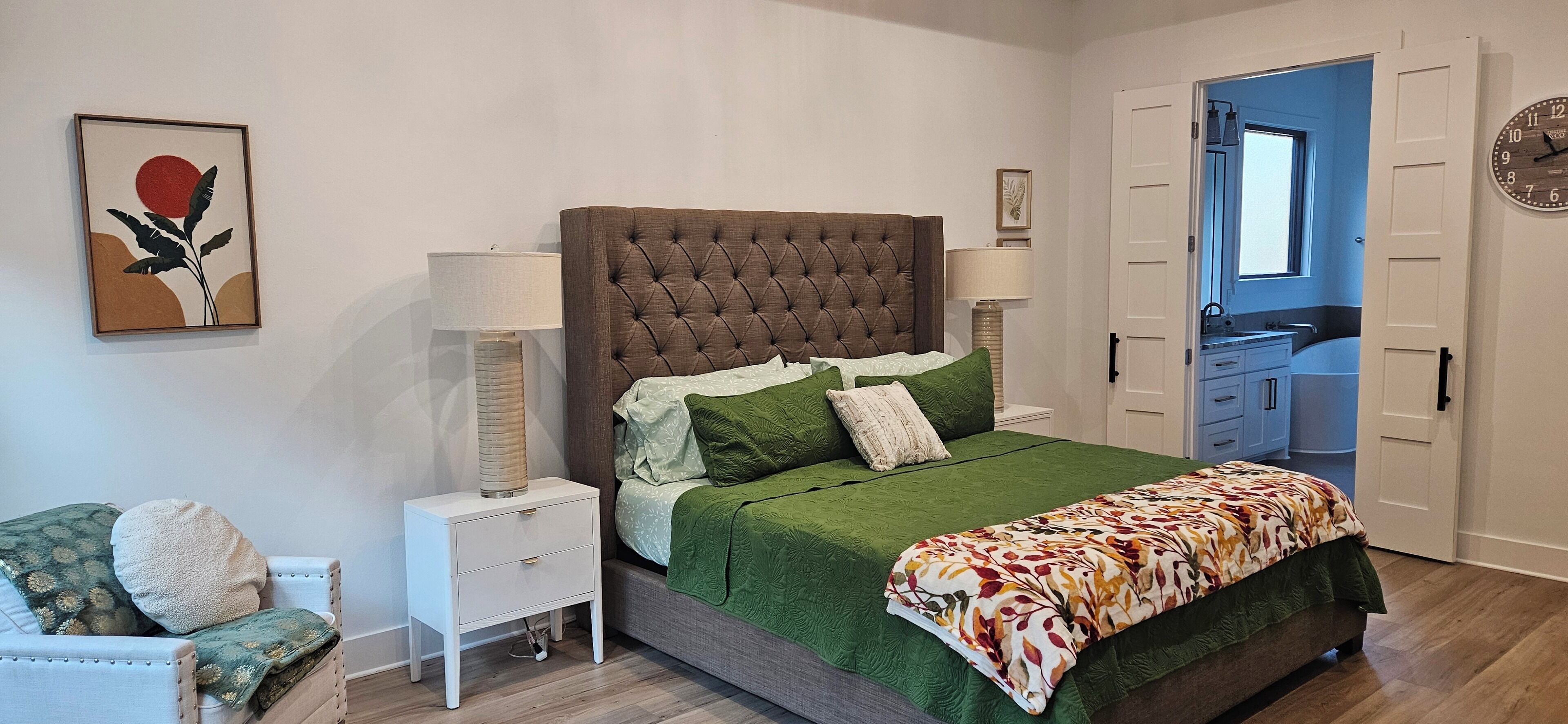 A bedroom with a tufted headboard, green bedding, a side table with lamps, a chair, and a door leading to a bathroom.