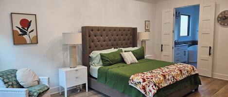 A bedroom with a tufted headboard, green bedding, a side table with lamps, a chair, and a door leading to a bathroom.