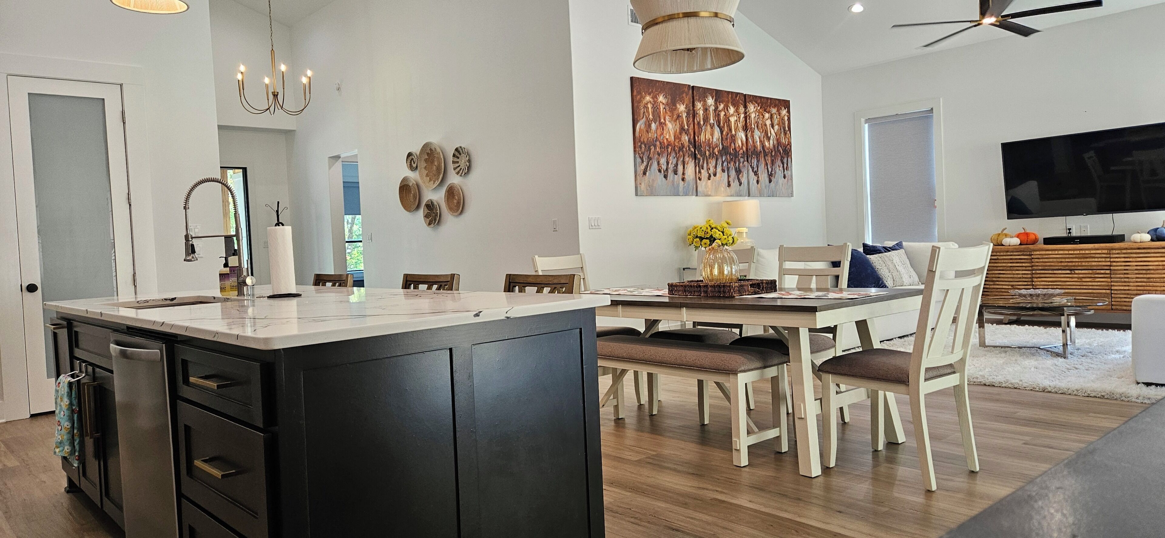 A modern kitchen with a central island, dining area with chairs, a large flat-screen TV, and decorative wall art.