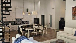 Un área de estar amplia con un sofá blanco, una mesa de comedor con sillas, una cocina con armarios blancos y una escalera.