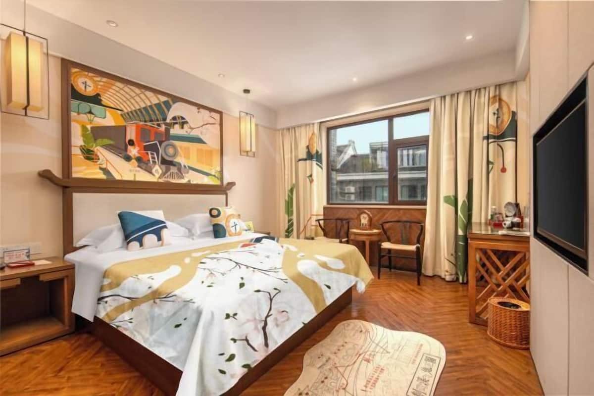 A hotel room with a bed, a desk, a chair, a window, and a painting on the wall.