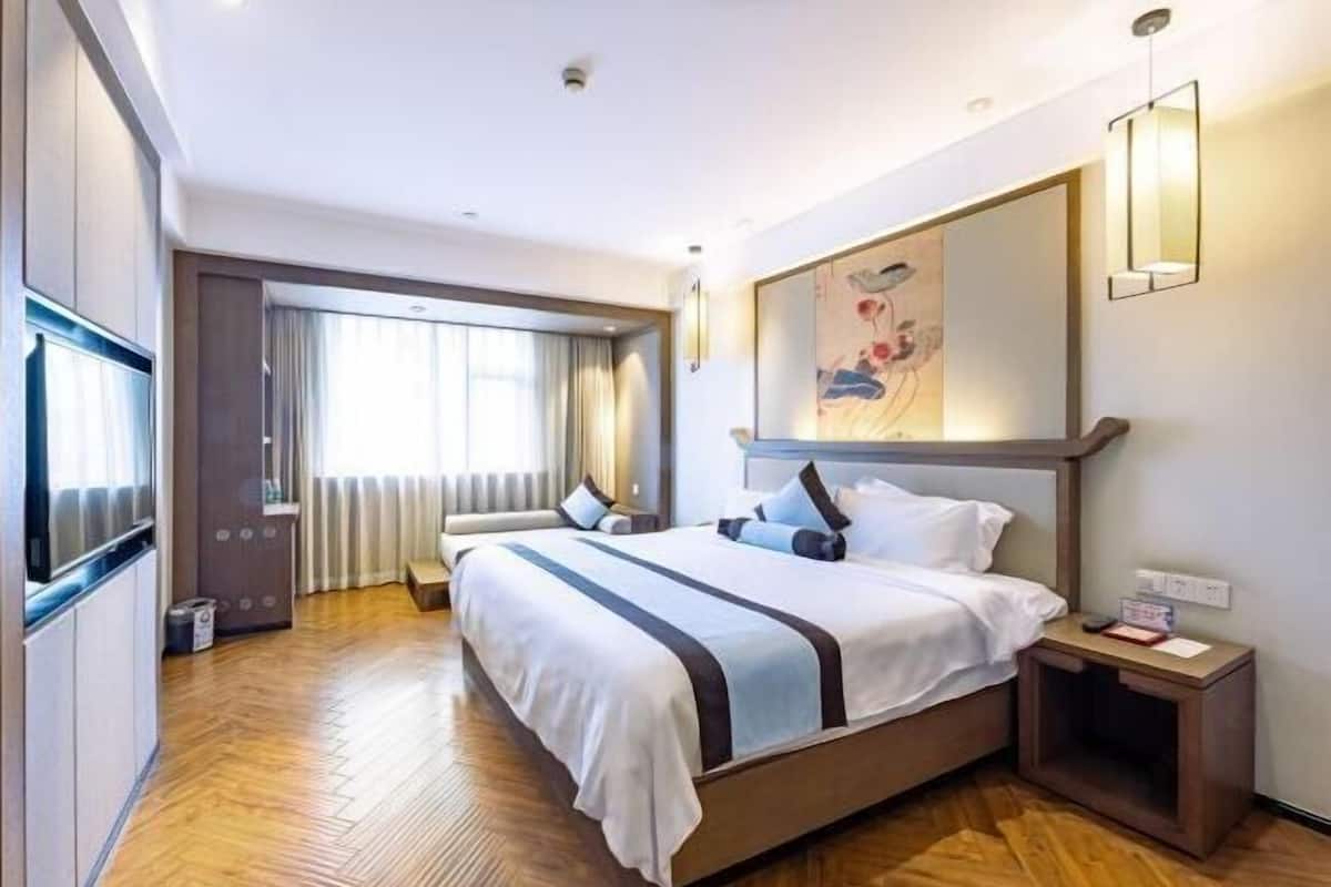 A modern hotel room with a large bed, a wooden floor, a large window with curtains, a painting on the wall, and a bedside table with a lamp.