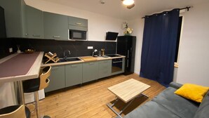 Une cuisine compacte équipée d’un four à micro-ondes, d’un évier et d’un ventilateur de plafond. Un canapé gris et une petite table en bois se trouvent dans l’espace salon.