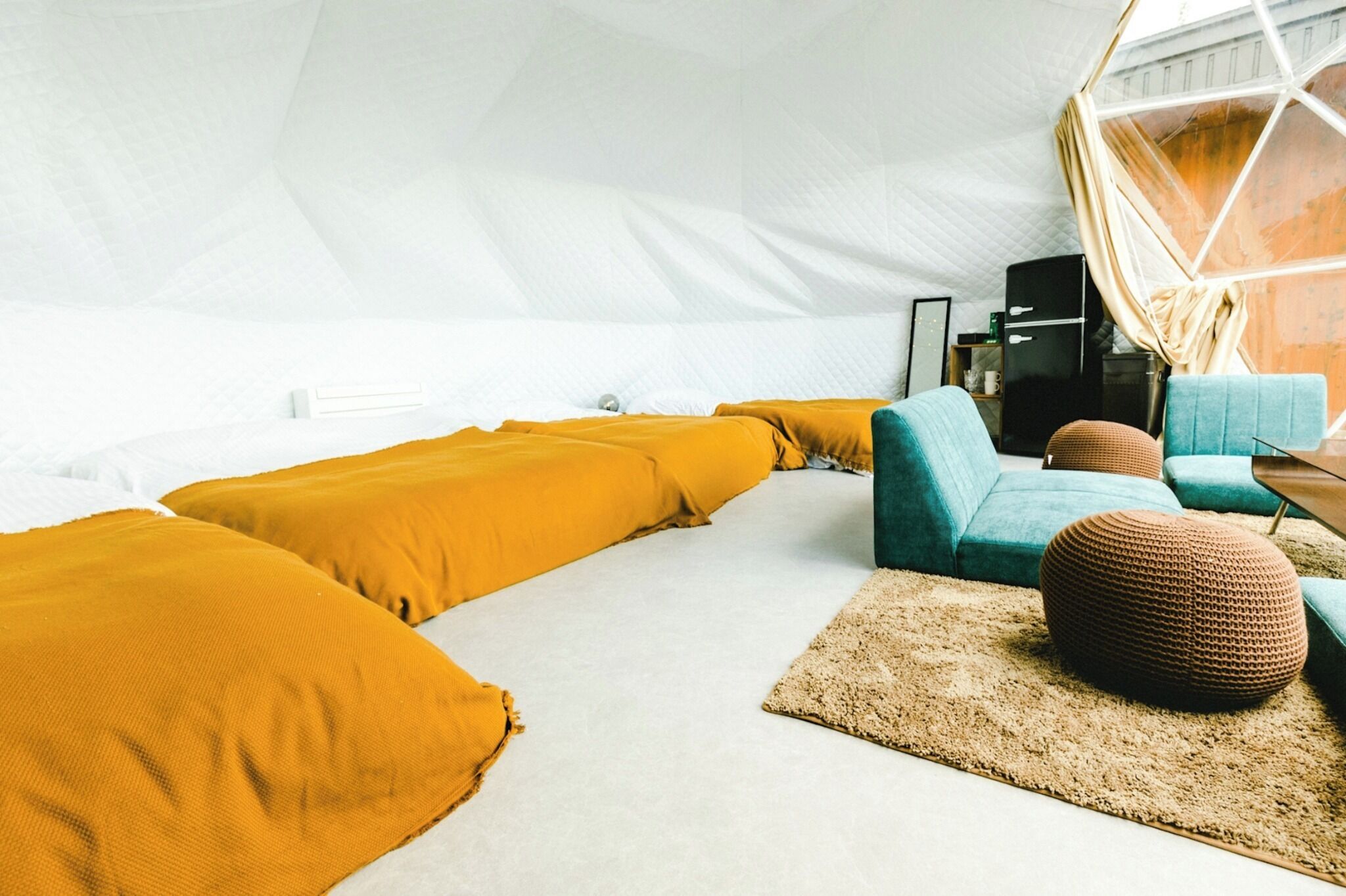 A tent-style room with a white interior, featuring a sofa set with mustard and teal cushions, a brown woven ottoman, and a wooden coffee table.