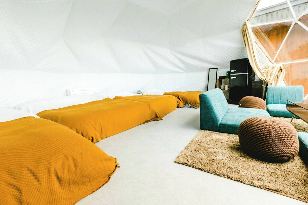 A tent-style room with a white interior, featuring a sofa set with mustard and teal cushions, a brown woven ottoman, and a wooden coffee table.