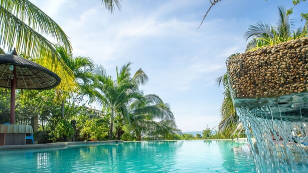 A clear swimming pool with a pebble-lined edge, surrounded by palm trees and a waterfall feature. - Spacious 5-bedroom Panglao Home with FREE transfers, Infinity Pool, WiFi, & A/C (Panglao)