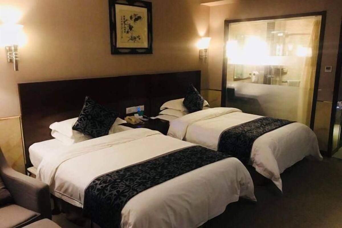 Une chambre d’hôtel avec deux lits, une table de chevet, une chaise et une salle de bain visible à travers une porte en verre.