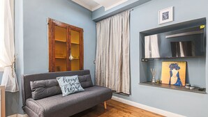 A room with a grey sofa, wooden floor, a wooden cabinet, a curtain, and a shelf with a painting. - Apartments Florence - Blue Serendipity (Firenze)