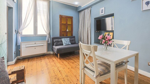 A room with a wooden floor, a grey sofa, a white table with chairs, a TV mounted on the wall, and a window with curtains. - Apartments Florence - Blue Serendipity (Firenze)