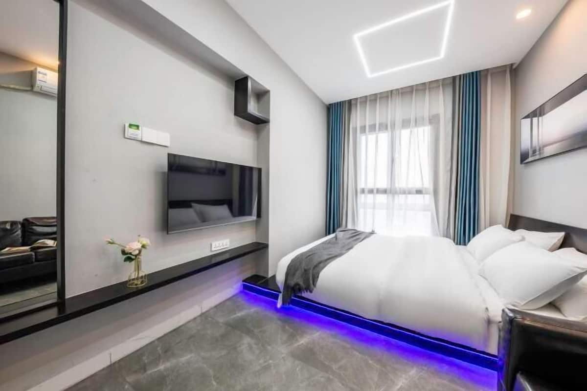 A modern hotel room with a bed, a flat-screen TV, a mirror, and a wall-mounted control panel.