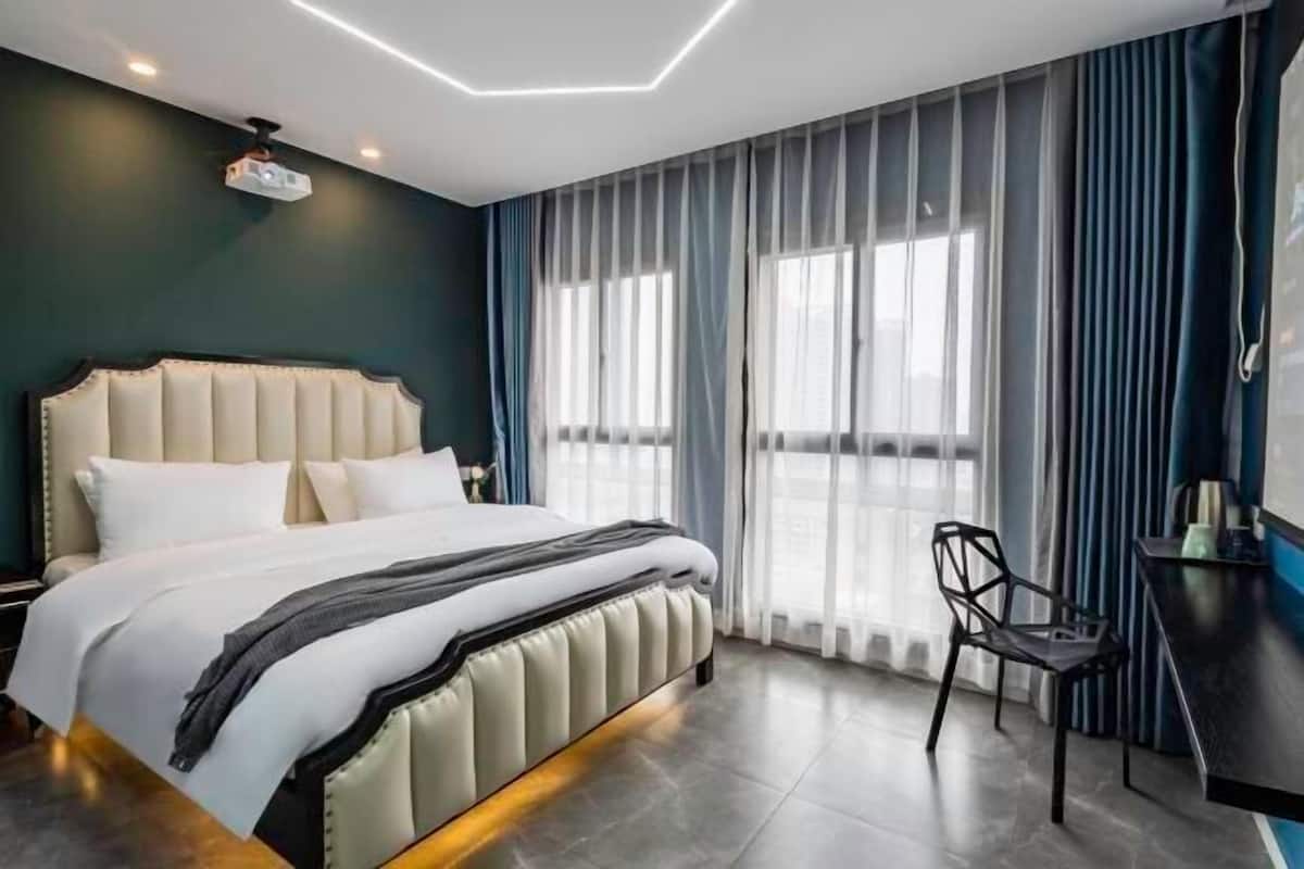 A modern hotel room with a large bed, a desk, and a view of the city through sheer curtains.