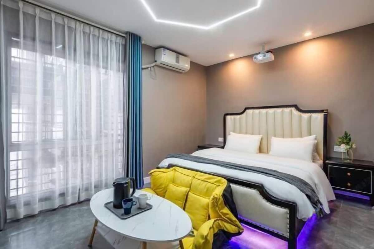 A hotel room with a bed, a bedside table with a cup and a tray, a projector on the ceiling, and an air conditioning unit.