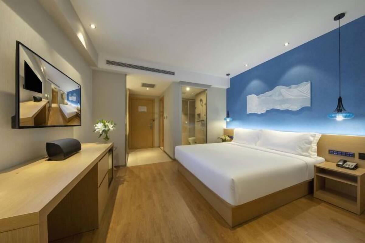 A modern hotel room with a large bed, bedside table, and a desk with a chair.
