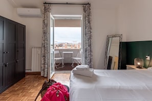 A hotel room with a bed, a wardrobe, a radiator, a window with curtains, a balcony with chairs, and a mirror.