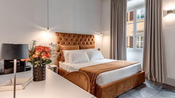 A hotel room with a tufted headboard, a bed with white linens, a bedside table with a lamp, and a vase with flowers.