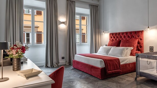 Een slaapkamer met een rood beklede hoofdeinde, een witte spreidekbed, een bureau met een lamp en een bloemenvaas.