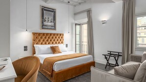 A hotel room with a tufted headboard, a bed with white and brown bedding, a small table, a chair, and a framed picture on the wall.