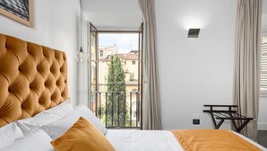 A hotel room with a tufted headboard, a bed with white and orange bedding, a view of buildings and trees through a window, and a small table with a chair.