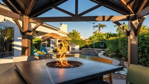 Un patio extérieur couvert, doté d’un foyer, de sièges et offrant une vue sur une piscine et un jardin.