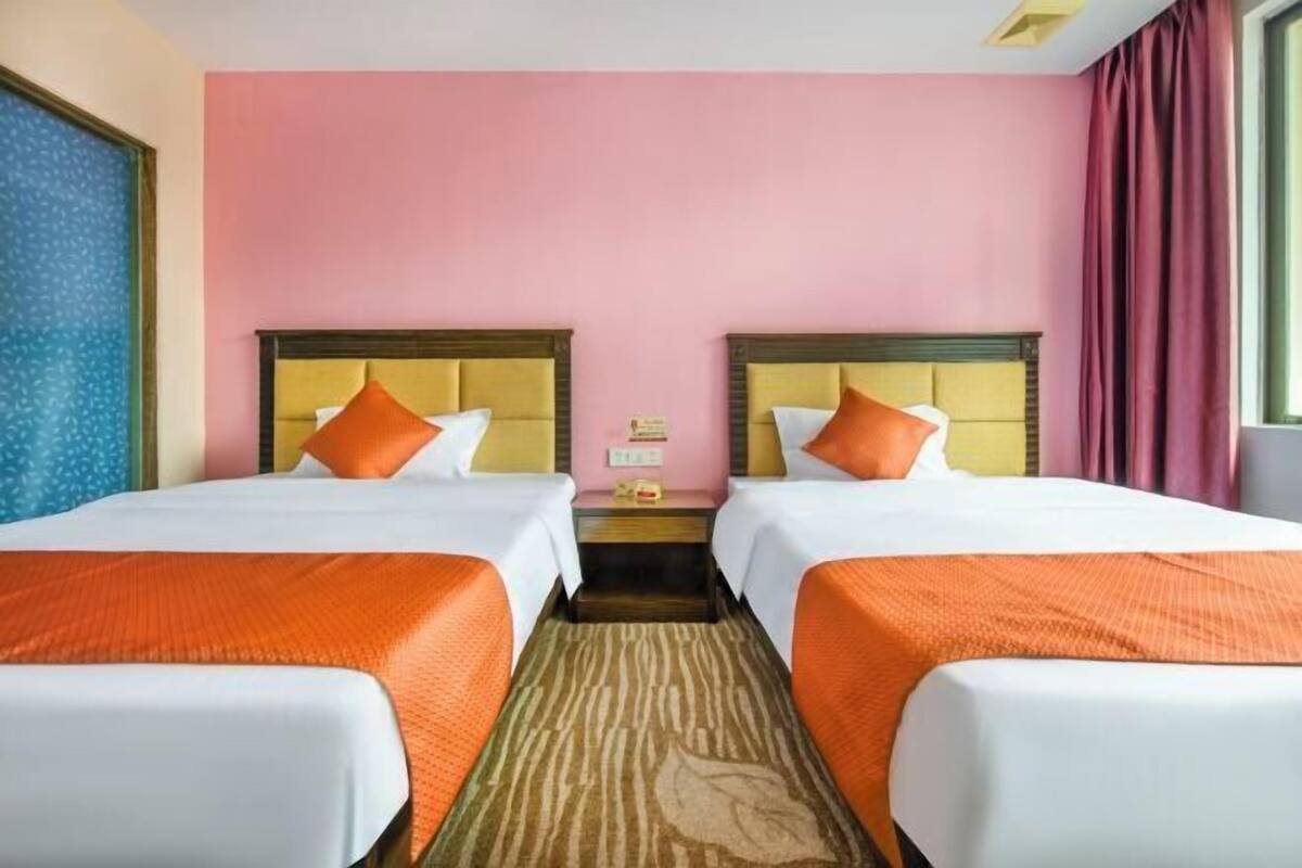 Camera d'albergo con due letti, ciascuno con testiera in legno, biancheria da letto bianca e cuscini decorativi arancioni. La stanza presenta una parete di colore rosa e una finestra con vista.
