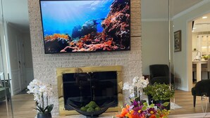 Een woonkamer met een flatscreen televisie aan een bakstenen muur, een open haard en een ronde salontafel met een afstandsbediening.