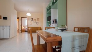 Un comedor con una mesa y sillas de madera, un mantel blanco y una estantería verde al fondo.