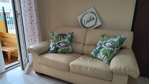Un sofá de cuero color beige con dos almohadones con estampado de hojas tropicales, un letrero en la pared con el nombre "Linda", y vista a una banca de madera a través de una puerta de vidrio.