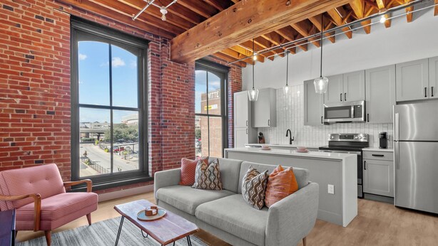 A modern kitchen with stainless steel appliances, a grey sofa with patterned cushions, and a large window offering a view of the city. - Riverfront DWTN Loft W/D Gated Parking (St. Louis)