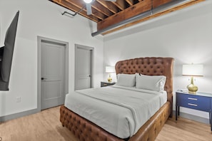 Une chambre à coucher avec une tête de lit capitonnée, un lit recouvert de linge de lit blanc, deux tables de chevet avec des lampes et un téléviseur fixé au mur.