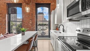 A modern kitchen with a brick wall, a large window, and a dining area with a white table and brown chairs. - Downtown Loft on Riverfront Next to Arch & Convention Center (St. Louis)