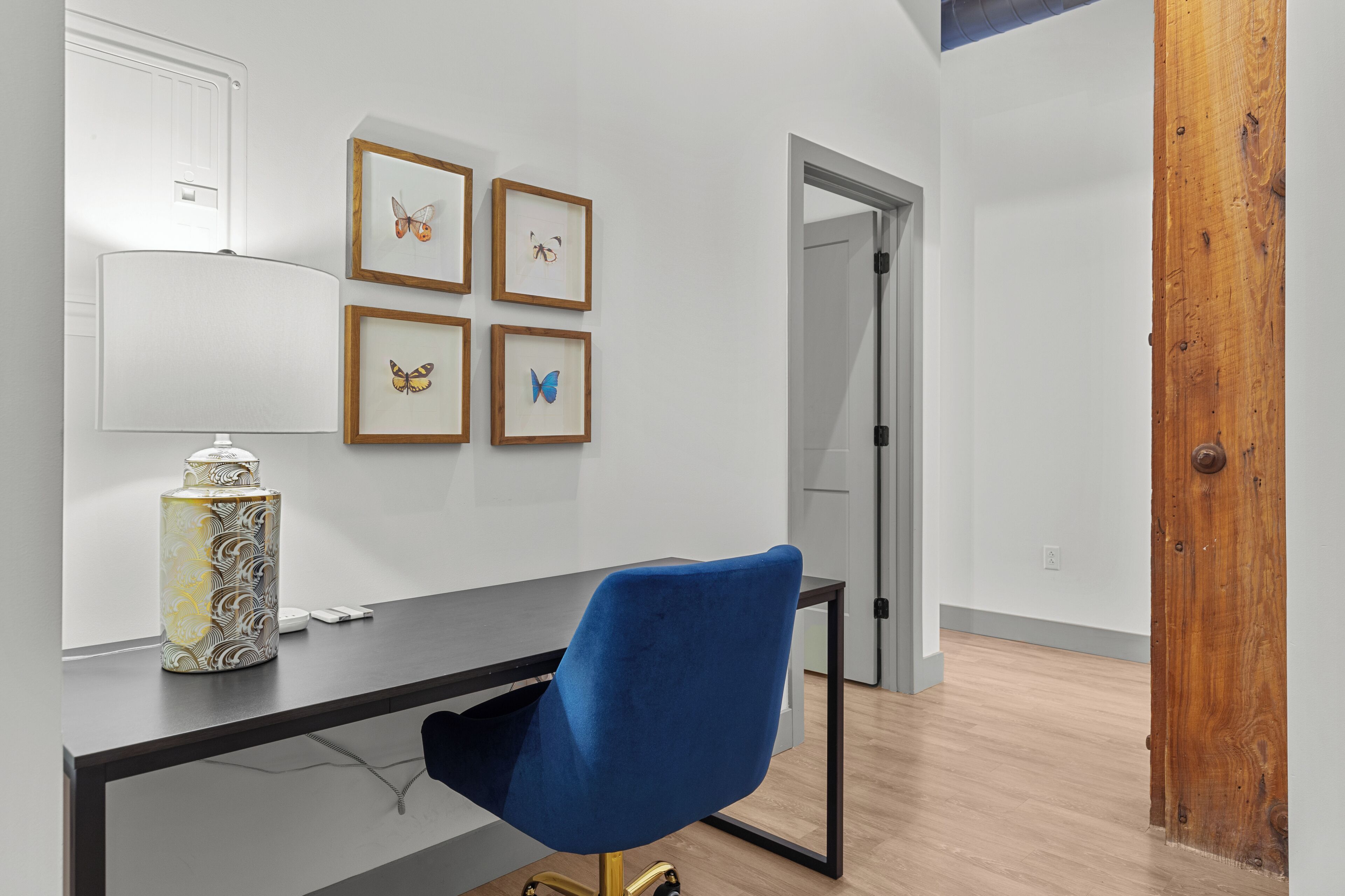 Ein moderner Arbeitsraum mit Schreibtisch, blauem Sessel, dekorativer Vase und gerahmten Schmetterlingsbildern an der Wand.