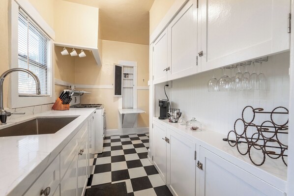 A narrow kitchen with white cabinets, a black and white checkered floor, and a coffee machine.