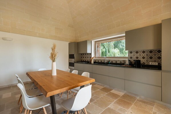 A modern kitchen with a wooden dining table and white chairs, a window with a view of greenery, and patterned backsplash tiles.