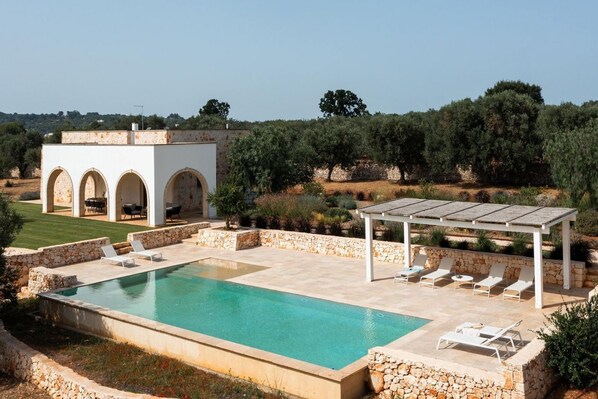 A swimming pool with lounge chairs, a pergola, and a white building with arches.