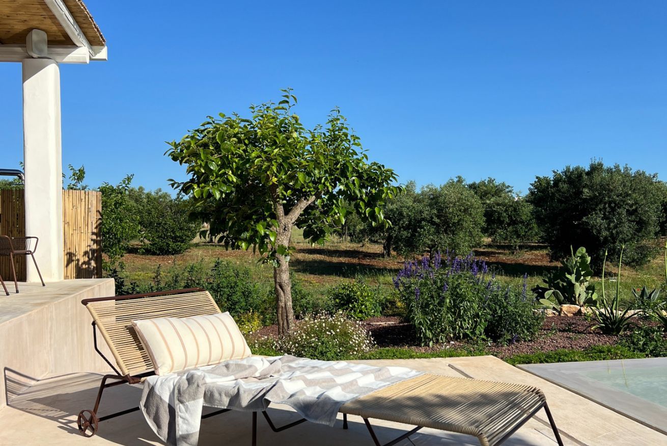 Ein sonnendurchfluteter Sitzbereich im Freien mit zwei Liegestühlen, einem kleinen Tisch und Blick auf Garten und Pool.