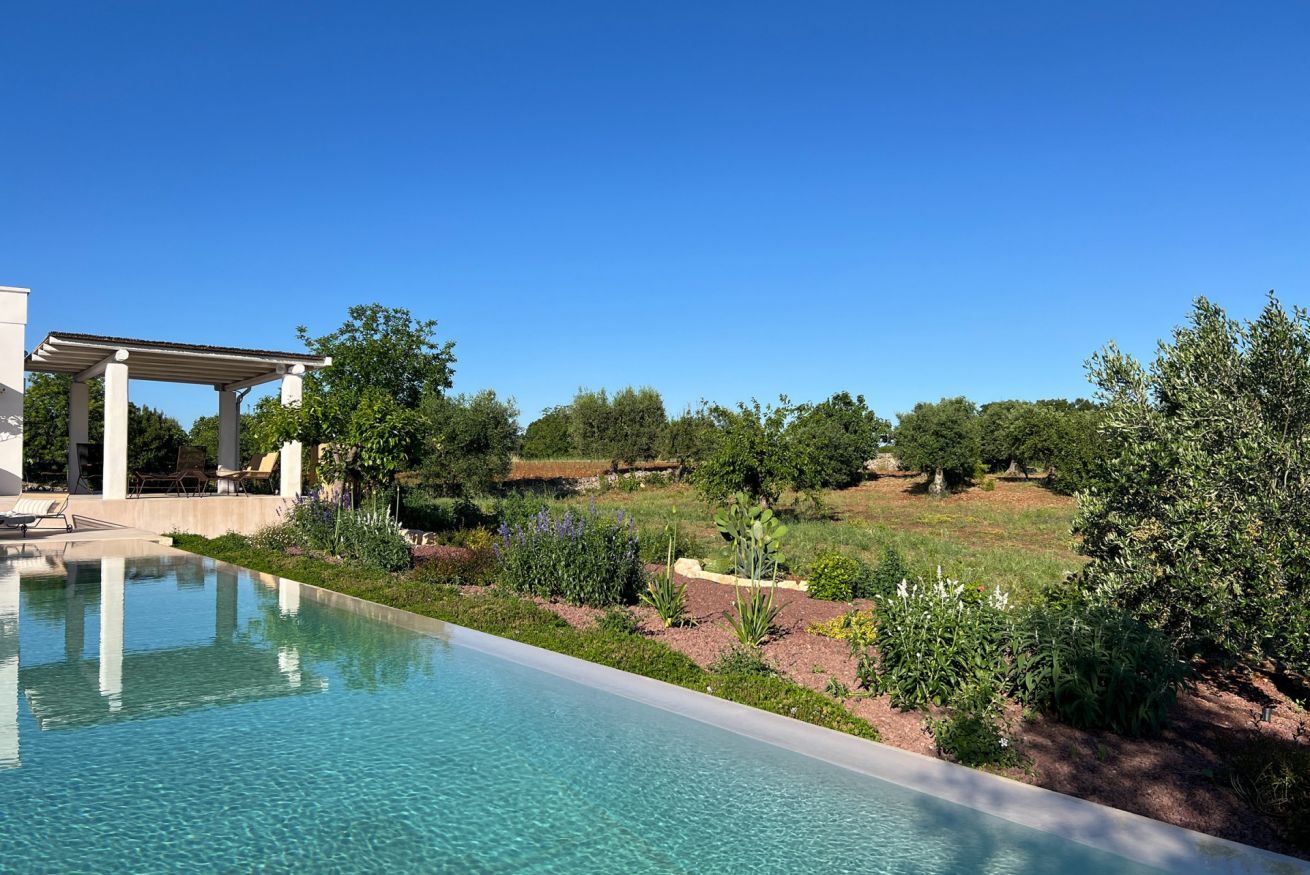 A clear swimming pool with a pergola and lounge chairs, surrounded by landscaped greenery and trees.