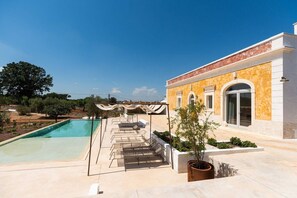 A pool area with lounge chairs and a pergola, surrounded by a landscaped garden and a building with arched windows.