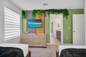 Une chambre avec un grand lit, une commode, un téléviseur fixé au mur, un papier peint à motifs tropicaux, et une vue sur la pièce adjacente.