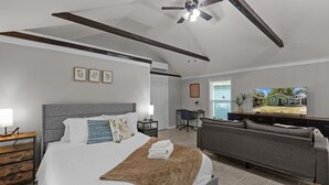 A bedroom with a bed, bedside tables, a desk, a chair, a grey sofa, a ceiling fan, and a TV displaying a house.