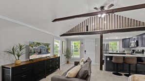A spacious living room with a grey sofa, a TV on a wooden cabinet, and a kitchen with a bar area.
