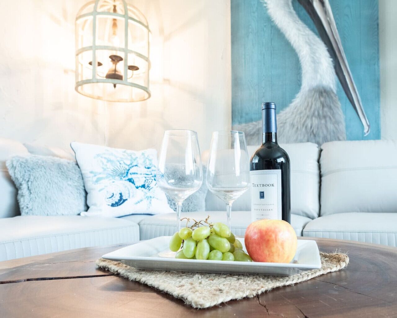 Una mesa de comedor con una botella de vino, dos copas de vino, un plato con uvas y una manzana, y una escultura decorativa de un pájaro al fondo.