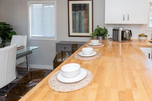 Cocina moderna con encimera de madera, armarios blancos y un comedor con mesa y sillas de vidrio.