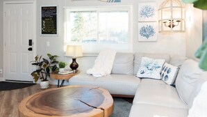 A living room with a round wooden coffee table, a beige sofa, a small side table with a lamp, and a door with a sign. - Cozy and Stylish-Walk To The Beach - Sleeps 5 (Grayland)